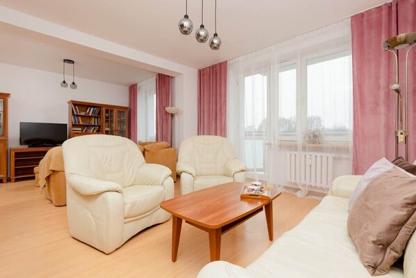 A spacious and well-lit living room featuring cream sofas, a wooden coffee table, and pink curtains, creating a cozy and elegant atmosphere.