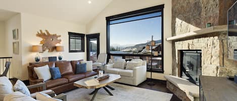 A modern living room featuring a large window with a scenic mountain view, a brown leather sofa, a white armchair, a wooden coffee table, and a stone fireplace.
