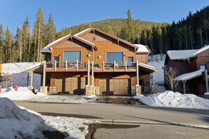 A duplex cabin with wood and stone exterior, two garages, and snow-covered surroundings is seen with tall pine trees and a forested hill in the background under a clear sky.