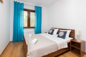 A bedroom featuring a large bed with a white bedspread and decorative blue pillows. Bright blue curtains hang by the window, and there are bedside tables with small lamps.