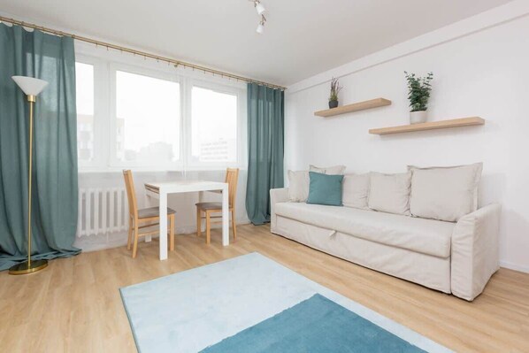 A cozy living room with a white sofa, a small coffee table, a potted plant, and large windows covered by turquoise curtains.
