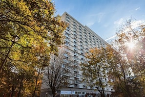 A view of a tall residential building surrounded by trees, bathed in sunlight. The mix of urban and natural elements creates a vibrant atmosphere.