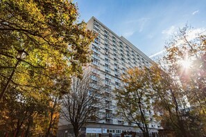 A view of a tall residential building surrounded by trees, bathed in sunlight. The mix of urban and natural elements creates a vibrant atmosphere.