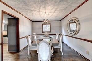 Elegant and modern dining room with a long wooden table, white chairs, chandelier, and mirror, offering a stylish and comfortable dining experience.