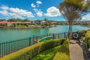 A beautifully landscaped waterfront backyard with lush hedges and a brick pathway leads to a private dock with a green kayak. Across the tranquil canal, charming homes line the shore under a bright blue sky, framed by gentle hills in the distance.