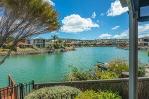 This peaceful waterfront view captures a serene canal lined with modern homes and private jetties. Lush greenery and vibrant landscaping frame the foreground, while a small boat rests gently at the water’s edge. Rolling hills rise in the distance under a 