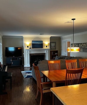 Reverse view of living and dining area with gas fireplace, 55" inch 4k Roku TV, book case filled with games and books for kids up to adults, shaker-style Amish table, piano, and vintage Marina sign.