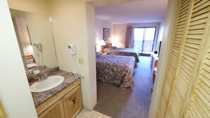 Hotel room with two beds, a television, a small table, and sliding glass doors leading to a balcony. In the foreground, there is a bathroom sink with a wall-mounted hairdryer.