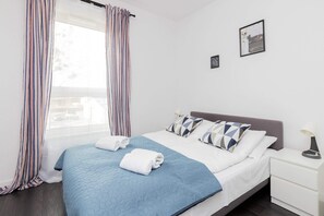 A bright bedroom with a neatly made double bed, white bedding, and a small bedside table.