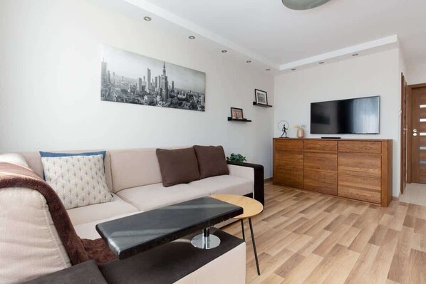 A cozy living room with a brown sectional sofa, coffee table, and a wall-mounted TV. The decor includes wall art and a soft carpet on the wooden floor.