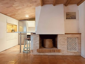 This spacious and modern kitchen features a unique fireplace. The kitchen also has a large refrigerator, a built-in cabinet, and a wooden ceiling with exposed beams. The warm terracotta tile floor adds a touch of traditional charm to the modern design.