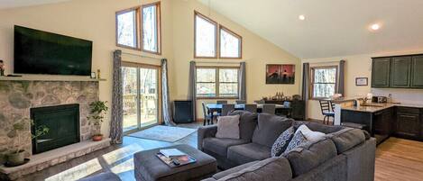 Living room with high vaulted ceiling, big windows letting in natural light, a stone fireplace and streaming TV.