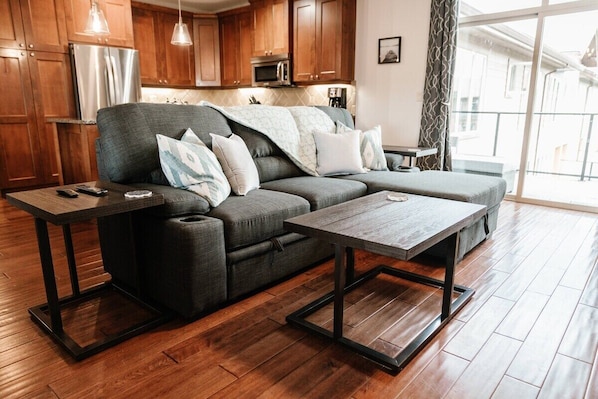 Unwind in a cozy living room featuring a spacious dark gray sectional, accented with soft white and blue pillows. Enjoy the warmth of hardwood floors, a stylish coffee table, and an inviting view of the modern kitchen with stainless steel appliances.