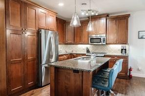 Cook and gather in a modern kitchen with a large island featuring dark granite countertops, wooden cabinets, and stainless steel appliances. Two bar stools offer seating under elegant pendant lights, making it a perfect spot for meals or mingling.