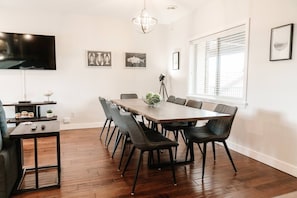 Gather around a sleek wooden dining table with black metal legs, seating ten comfortably in chic black chairs. Natural light pours in through a large window, highlighting the sea-inspired artwork and modern lighting above.