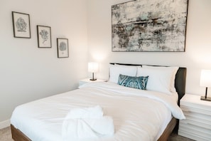 Relax in a serene bedroom featuring a plush bed with crisp white linens, accented by a teal-patterned pillow. Flanked by elegant nightstands and soft lighting, the space is adorned with abstract and floral art for a touch of color and style.