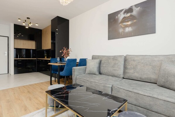 A modern living room featuring a gray sectional sofa, a glass coffee table, and a sleek black-and-wood kitchen in the background.

