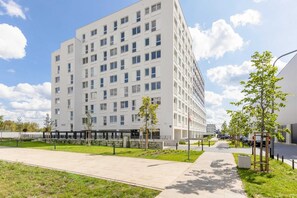A newly built residential building surrounded by green areas and pathways, set against a bright blue sky with clouds.