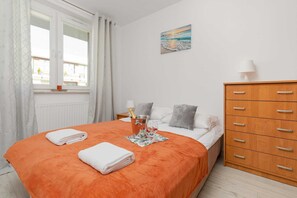 A bedroom with an orange bedspread, neatly folded towels, a window with curtains, a dresser, and artwork above the bed.