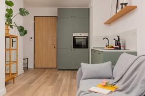 A living room corner with a small gray sofa, a wooden table, a dining area, and a plant, blending modern and natural design elements.