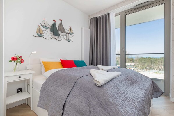 A cozy bedroom featuring a double bed with neutral bedding, decorative pillows, and nautical-themed artwork on the wall. Large windows allow plenty of natural light.