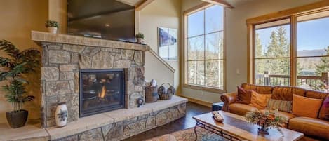 The spacious living area is anchored by a stunning stone hearth and fireplace with a large, flat-screen smart TV and bathed in natural light from the surrounding windows, offering tranquil nature views.