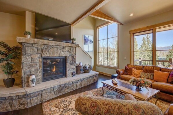 The spacious living area is anchored by a stunning stone hearth and fireplace with a large, flat-screen smart TV and bathed in natural light from the surrounding windows, offering tranquil nature views.