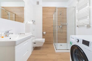 Bright and modern bathroom featuring a shower, sink, toilet, washing machine and wooden accents. The space is clean and functional.
