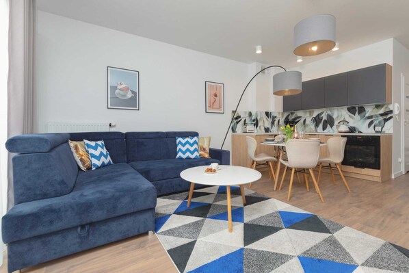 Modern living room featuring a cozy fold-out sofa, TV and kitchenette. The large window allows plenty of natural light to brighten the space.