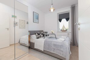 A stylish bedroom with a queen-sized bed, light gray bedding, and colorful accent pillows. There’s a simple nightstand and a wall-mounted lamp.