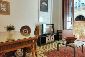 The living area features a large, glass-topped dining table surrounded by wicker chairs. There's a flat-screen television mounted on a black stand with wicker storage baskets.  The  French doors open to a balcony, providing a glimpse into the vibrant stre