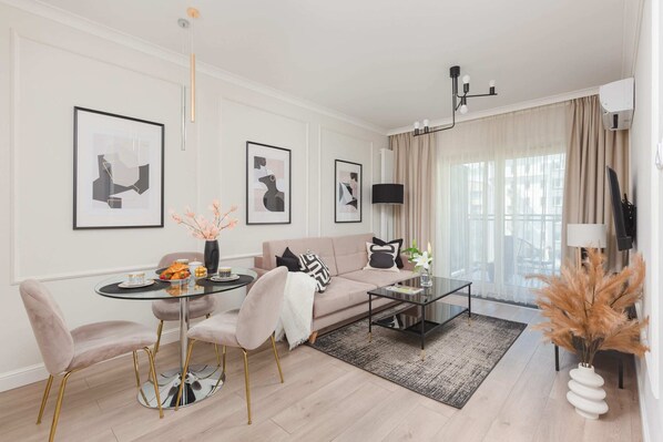 A well-lit living room with a beige sofa, decorative pillows, a glass coffee table with flowers, and modern wall art. The room is bright and styled with contemporary decor.