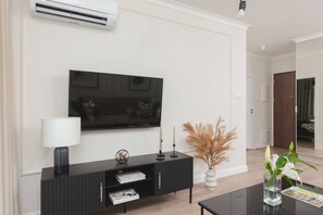 A corner view of a living area featuring a wall-mounted flat-screen TV, a sleek black console table with a lamp and decorative items. The space looks clean and minimalist.