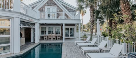 Poolside view of the home facing the back of the home. Four white chaise lounge sunning chairs on the right with umbrella. An exterior dining table under a cover by the kitchen.