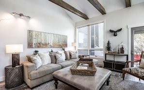 The living room in this home has it all - a roomy sectional sofa, oversized coffee table/ottoman, two reading chairs, a large flat-screen TV, gas fireplace with stone work, natural light, tree views and stylish wood beams.