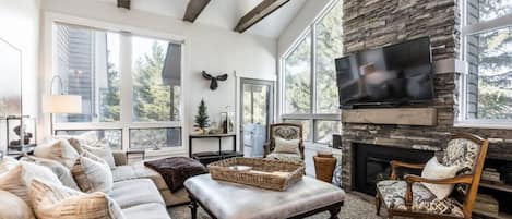 The living room in this home has it all - a roomy sectional sofa, oversized coffee table/ottoman, two reading chairs, a large flat-screen TV, gas fireplace with stone work, natural light, tree views and stylish wood beams.
