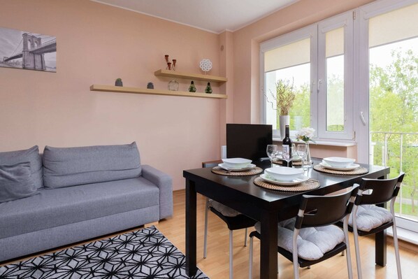 Cozy living and dining area with a gray sofa, wooden table, and bright natural lighting from large windows.

