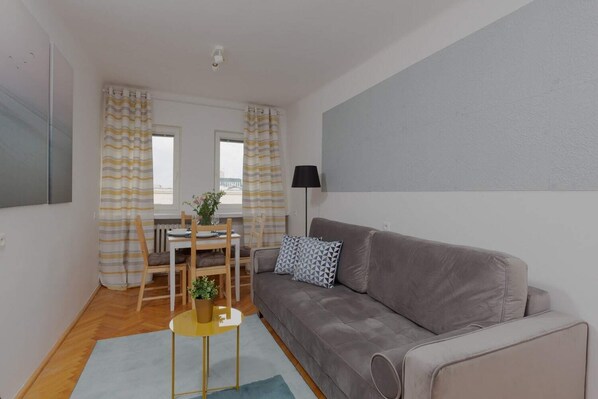Living room with a gray sofa, wooden table, and bright natural light coming through the windows.
