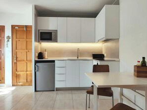 The bright and modern kitchen boasts sleek white cabinets and a compact layout. A small refrigerator, microwave, and coffee machine offer convenience for preparing meals, while a dining table and chairs provide a comfortable space for enjoying them. Natur
