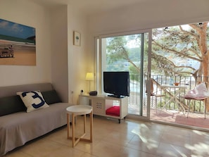 The living area features a comfortable sofa bed, a flat-screen television, and a small, stylish side table. The large window provides a beautiful view of the surrounding trees and the balcony beyond. It is a very luminous space.