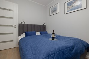 A bedroom with a double bed featuring a blue quilt and matching pillows. The room includes white storage furniture and minimal wall art.