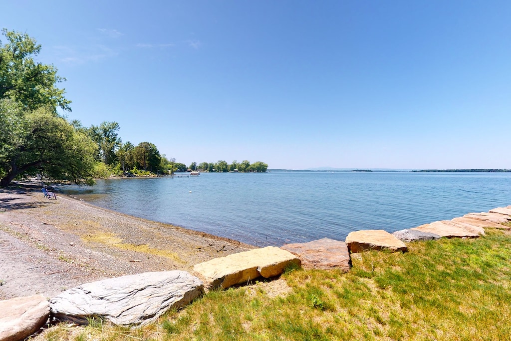 A rocky beach on Lake Champlain has grass on one side and several trees on the other under clear blue skies