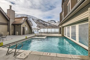 Outdoor pool next to a building with snowy mountains in the background. The pool area is clean and features safety signs, a metal handrail, and seating arrangements.