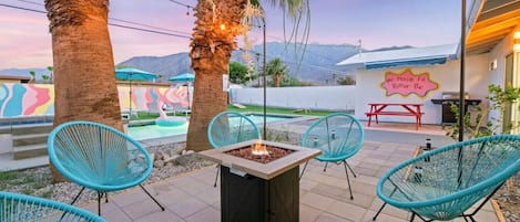 The fire pit is nestled in the corner of the pool, providing a cozy spot for conversation and relaxation. Warm, flickering flames dance and crackle in the night air. The light from the fire casts an orange glow on the pool and the surrounding area.