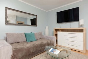 A functional living room with a sofa bed with soft cushions, a large mirror hangs on the wall above the sofa and on the other side there is a flat-screen TV on the wall.