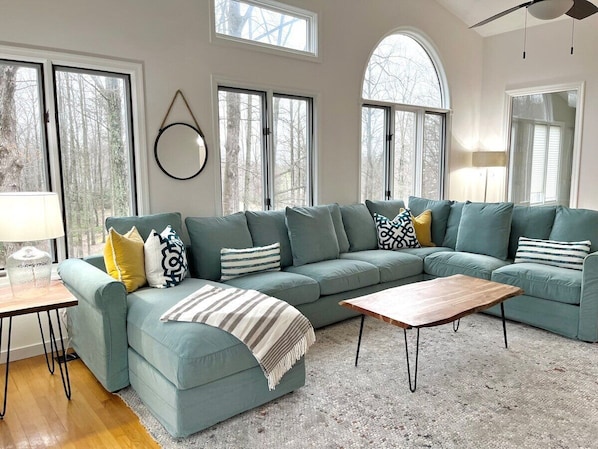 Main House: Second Floor- Large sectional sofa and picture windows in the upstairs living room create a great gathering space. Enjoy peaceful wooded/mountain views, patio doors for deck access, and a 65" Smart TV