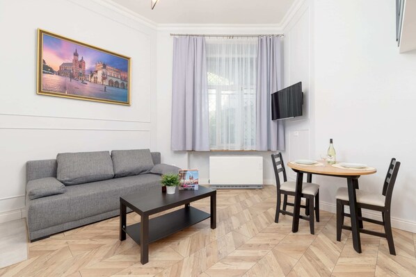 Living rooing room with a gray sofa, a small black coffee table, and a table with two chairs. A wall-mounted TV and framed artwork are visible.m 