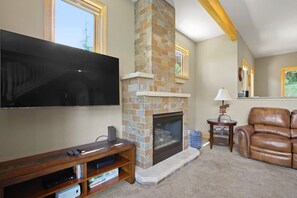 The spacious living room features wood beam accents, abundant natural light, two leather couches, a roomy leather armchair with an ottoman, coffee table, end table, lamps, a flat-screen TV and a striking, stone fireplace.