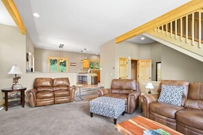 The spacious living room features wood beam accents, abundant natural light, two leather couches, a roomy leather armchair with an ottoman, coffee table, end table, lamps, a flat-screen TV and a striking, stone fireplace.