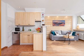 A bright and modern living space featuring a kitchenette with light wooden cabinets, a blue sofa, and a dining table for four. Large windows let in plenty of natural light.